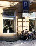 Wejście do dziennego domu. Widok z zewnątrz. Szklane drzwi wejściowe i trzy okna na kremowej fasadzie budynku. W oknach kwiaty doniczkowe. Przed budynkiem rower i znak drogowy "Parking"